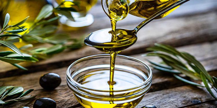 Pouring extra virgin olive oil from a spoon to a glass container. Olive branches comes from the left and right. Some olive oil bottles are out of focus at background. Black olives are on the table and complete the composition. The composition is on a rustic wooden kitchen table. Predominant colors are gold, green and brown. High resolution 42Mp studio digital capture taken with Sony A7rII and Sony FE 90mm f2.8 macro G OSS lens